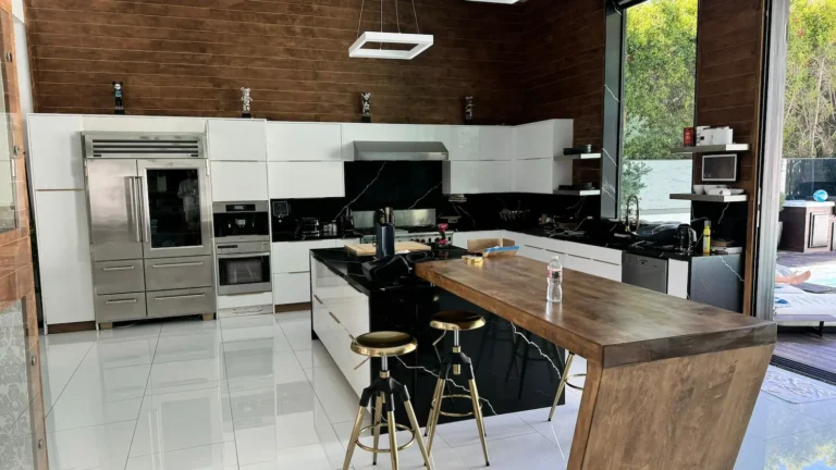 A high-end kitchen by Sean Design featuring a large black marble island with an attached cantilevered wood breakfast bar, white high-gloss cabinetry, and a dark wood-paneled wall. The space includes professional-grade stainless steel appliances and gold-accented barstools.