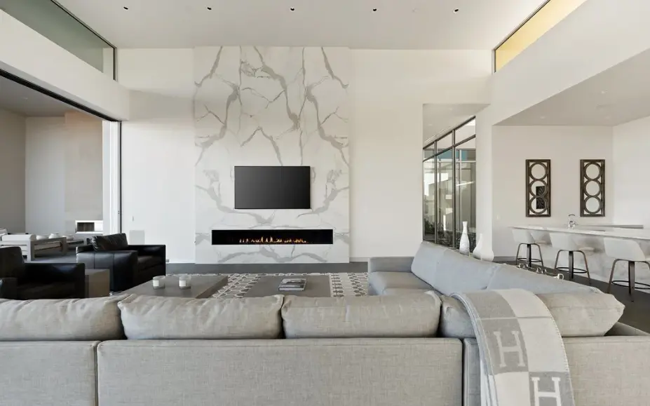 A spacious, bright living room featuring a massive white marble fireplace wall with grey veining, a mounted television, and a linear gas fireplace. The room includes a large grey sectional sofa, black leather armchairs, and a modern kitchen island with white barstools in the background.